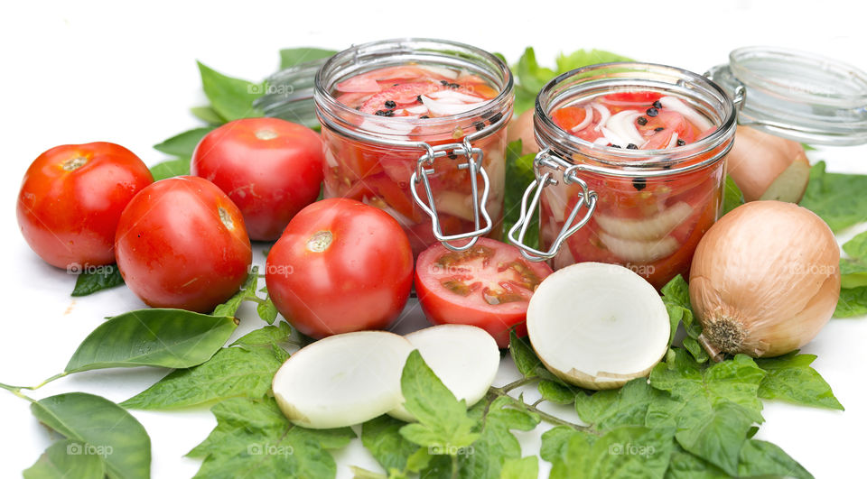 Whole tomatoes and  sliced onions and two glass  jars of pickled tomatoes and onions with peppercorns against white.