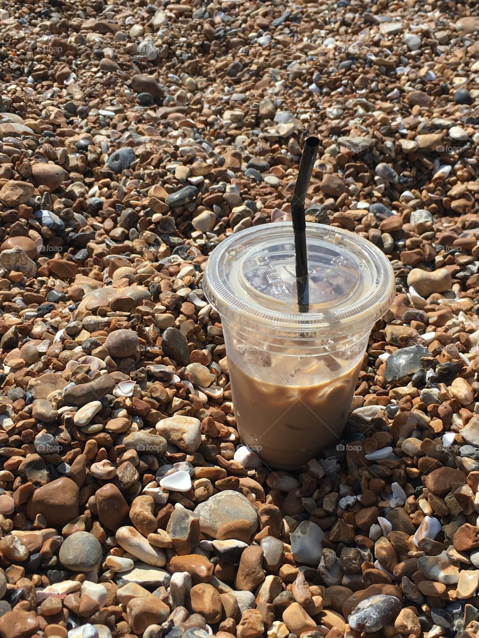 Half an iced latte in a plastic cup nestled in the rocks on a rocky beach 