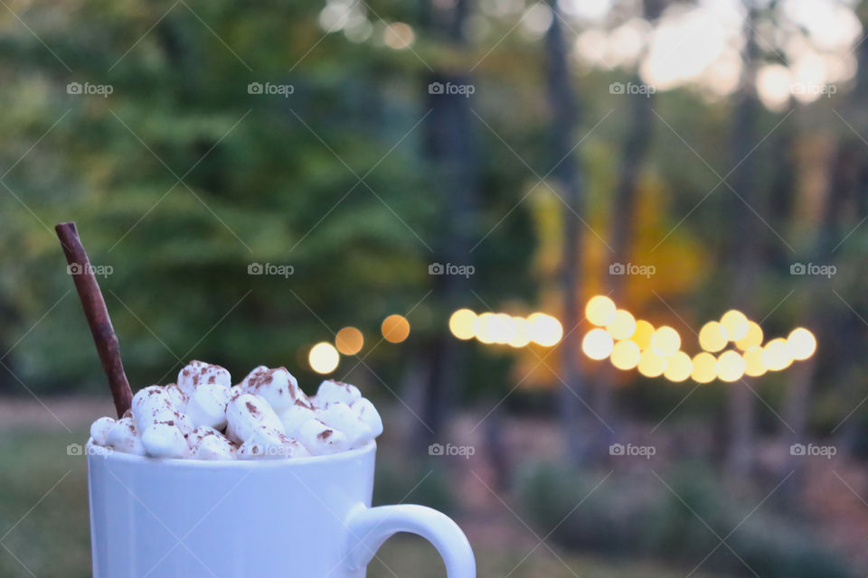 Hot drink with cinnamon stick and marshmallows on deck with lighted fire pit in the distance. 