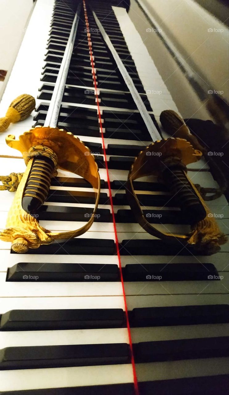 Reflection from the surface of the piano.  On the keys of the piano lies the sword of an Italian air force officer.  In the foreground there is a golden sword hilt, which turns into a metal blade.  The sword is reflected from the black polished surfa