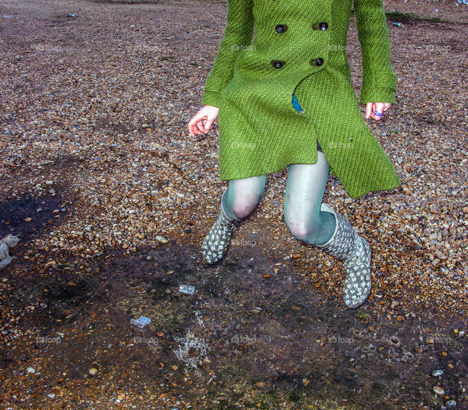 Woman in green coat jumps in puddles on a stony beach - Hastings UK 