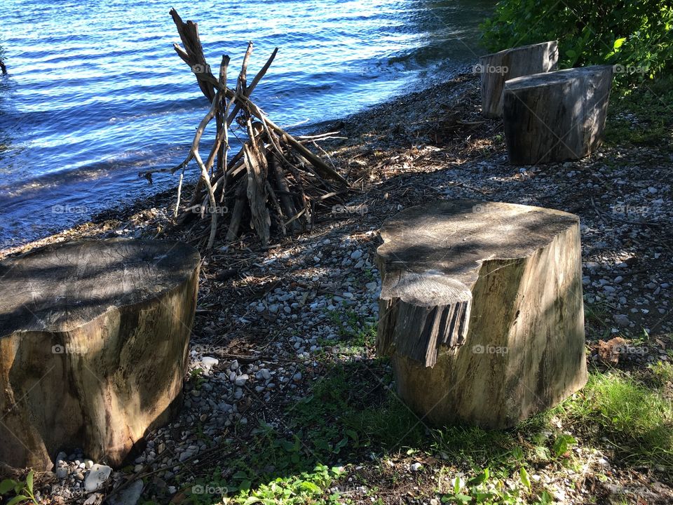 Stools made of cut wood logs scattered symmetrically around stack of wood ready for a summer campfire by the water, conceptual summertime vacation and relaxation off the beaten track photography