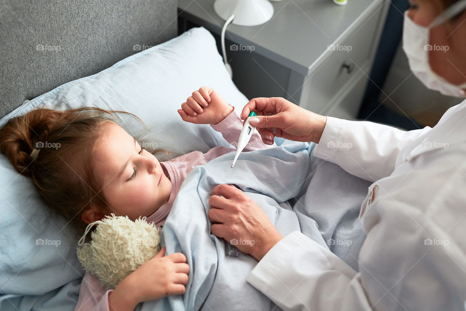 Doctor visiting little patient at home. Measuring the temperature of sick girl lying in bed. Woman wearing uniform and face mask. Medical treatment