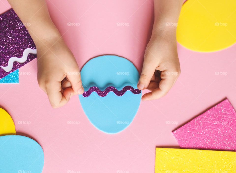 The hands of a little caucasian girl glue a lilac felt zigzag on a blue easter egg chile at a children table with a pink background, flat lay close-up. Concept diy, crafts, children needlework, Easter preparation.
