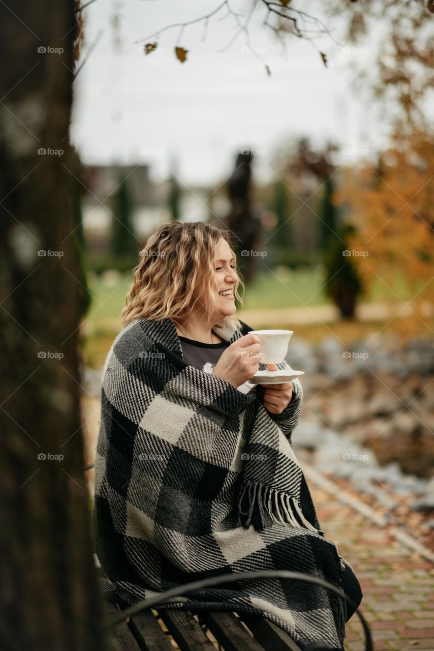 original autumn portrait of a young woman drinking tea wrapped in a plaid in an autumn park