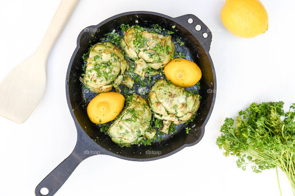 Flat lay of roasted artichokes with lemon and parsley in a cast iron skillet on a white table