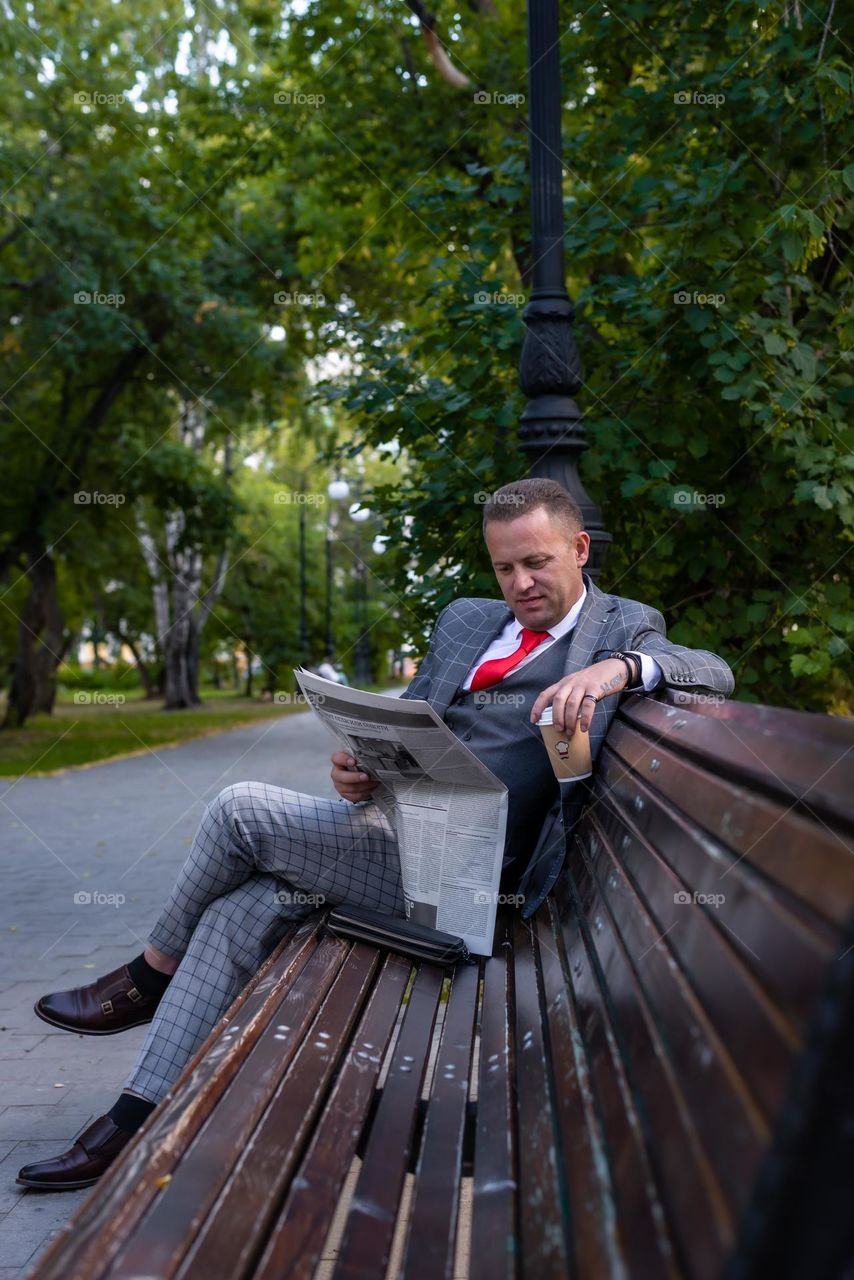 portrait of a man waiting for a meeting in a city park sits on a bench reading a newspaper