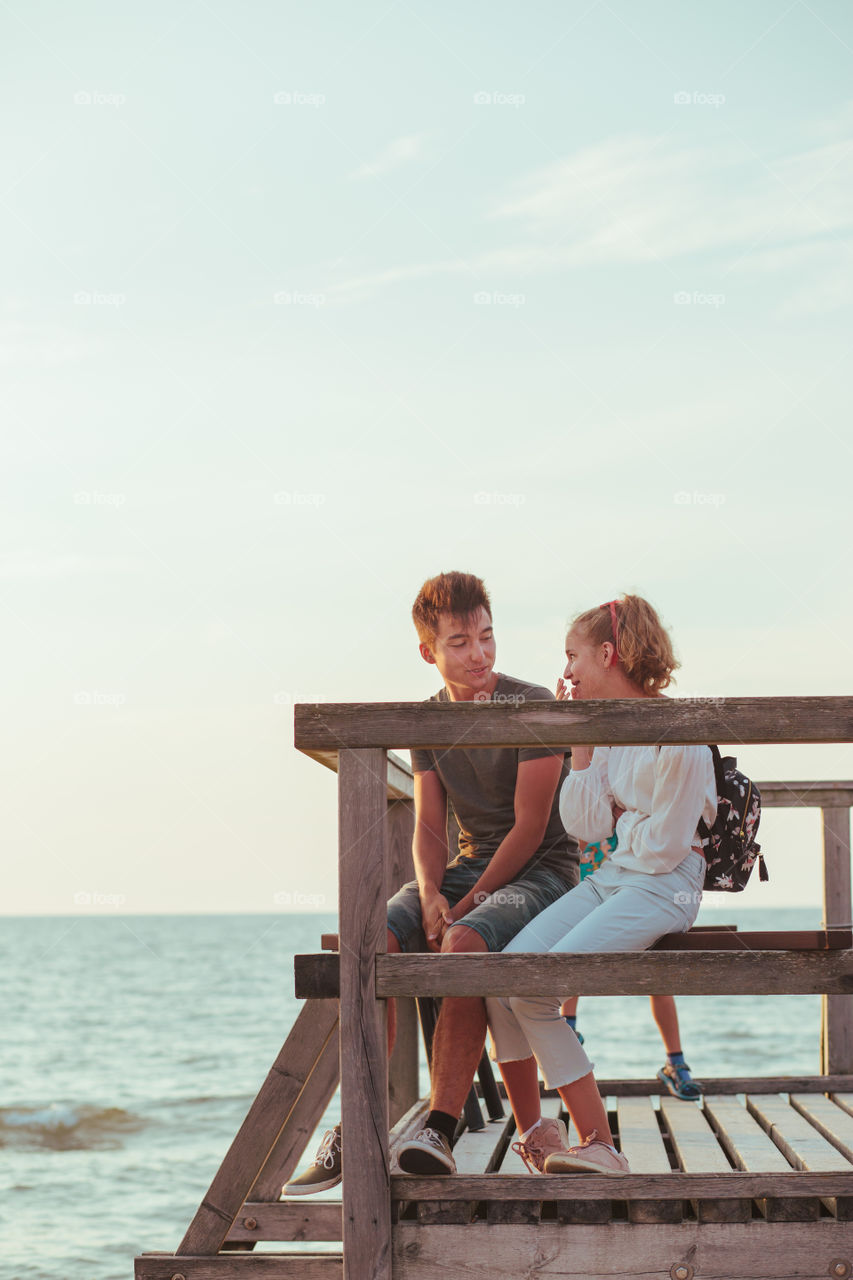 Happy smiling couple of young woman and man sitting on a pier over the sea during summer vacations. Copy space room for text