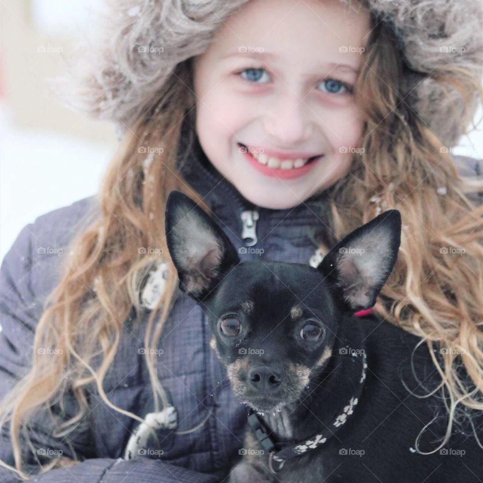A sweet little blue-eyed girl in a faux-fur jacket holding onto her precious little Chihuahua puppy. 