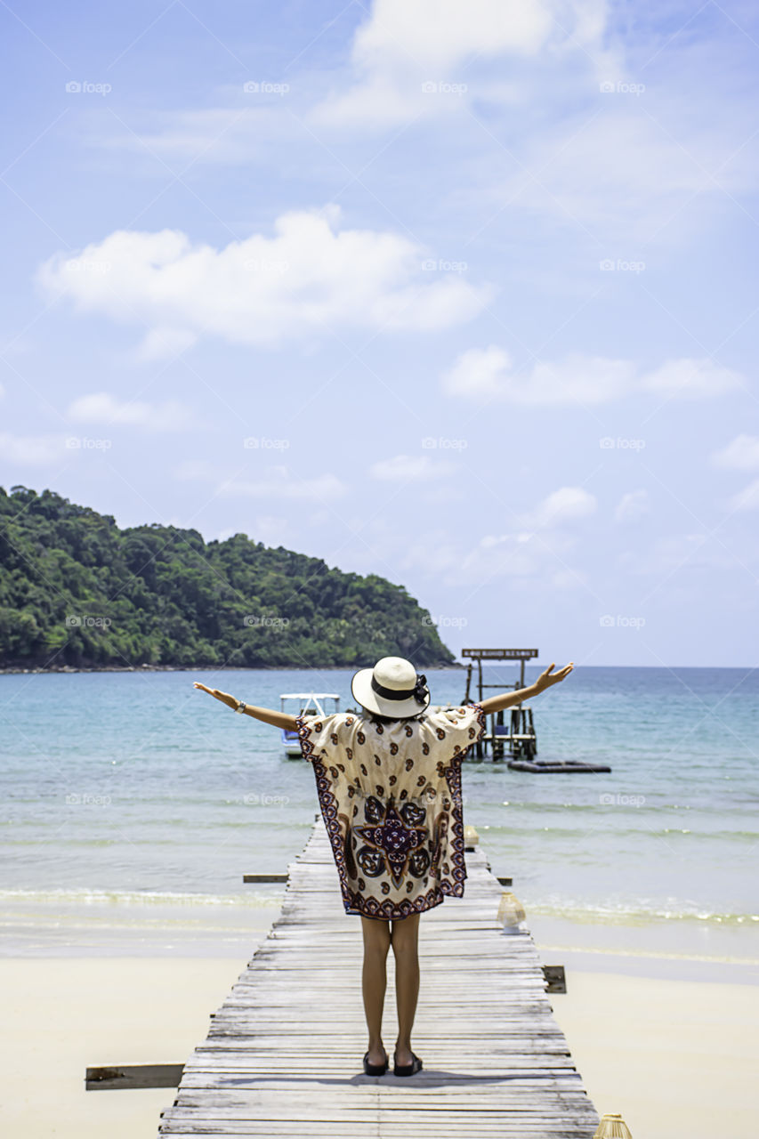 Women raise their arms and  wear a hat on the wooden bridge pier boat in the sea and the bright sky at Koh Kood, Trat in Thailand.