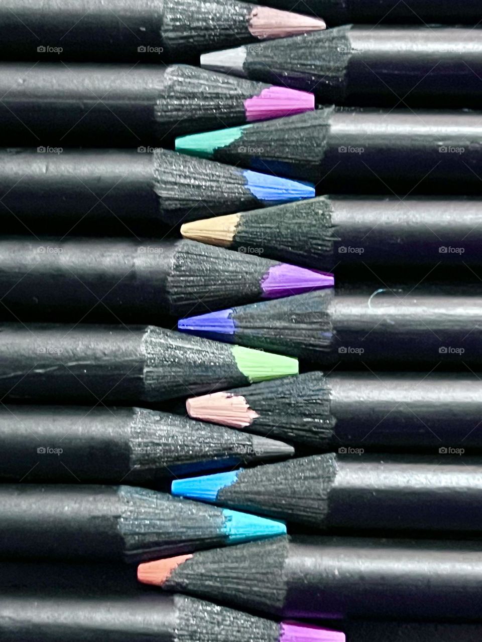 Imagem em close-up de lápis de cor preta com pontas de cores vibrantes, dispostos ordenadamente em fileiras paralelas, exibindo ferramentas artísticas e educacionais com foco na criatividade e organização.