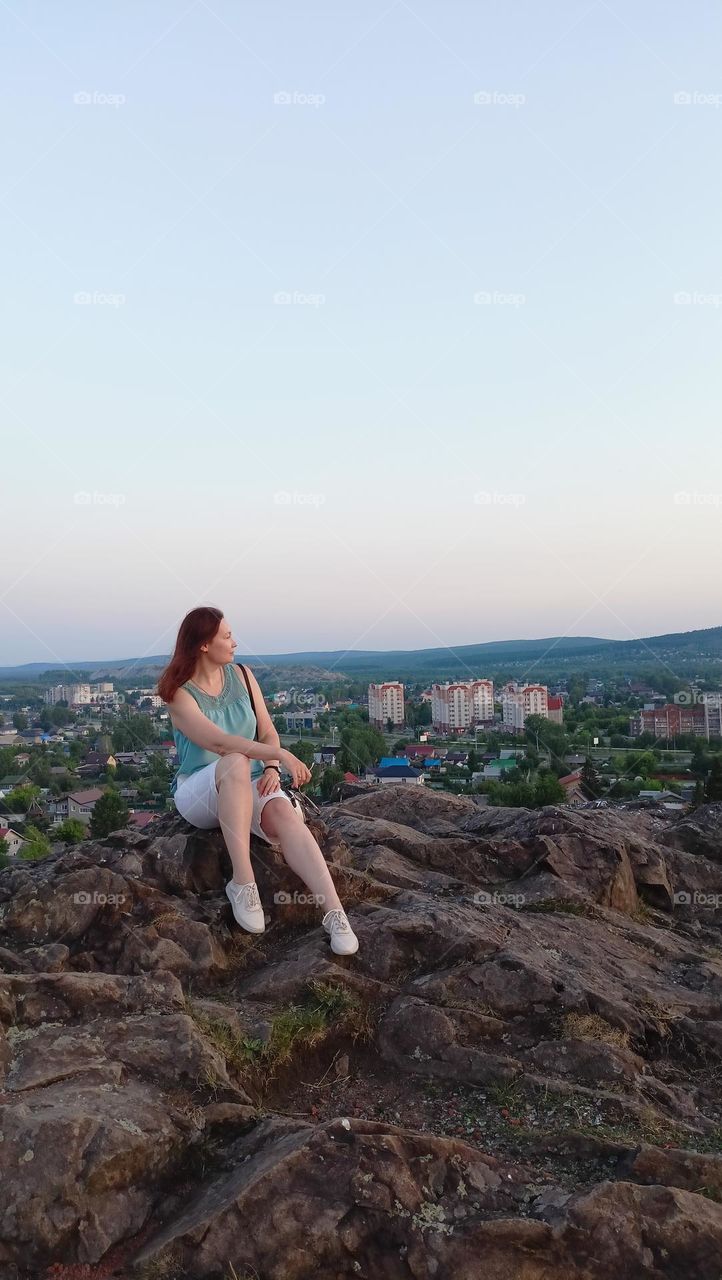 summer in the city, top view, urban landscape at sunset, a girl sits on a stone, looking into the distance, sky, mountains, city, woman in shorts