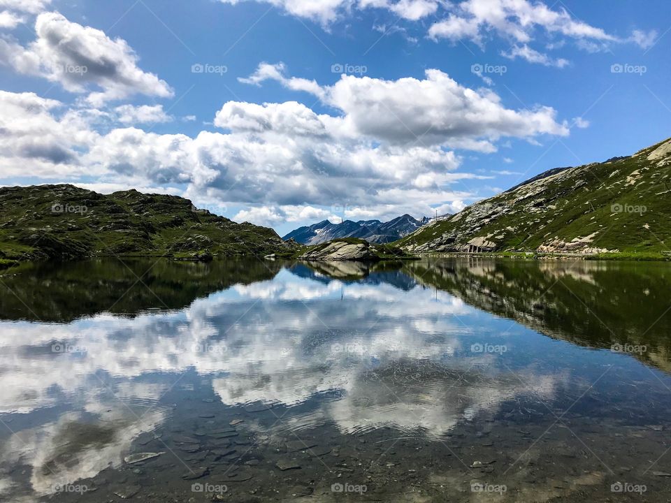 a lake in the middle of nature, with mountains surrounding it, clear water visible can on the bottom of the lake. in the blue sky there are large white clouds, color reflection into the water looks amazing