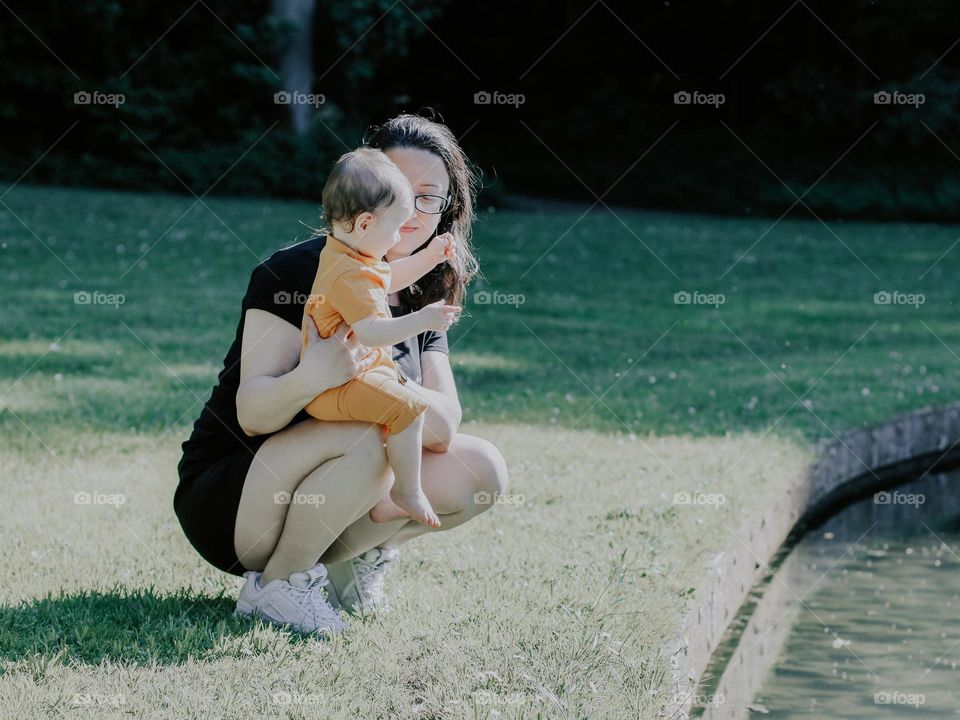 Portrait of a young caucasian brunette girl squatting holding her little baby daughter on her knees and looking tenderly at her in a park on a green lawn next to a pond,side view cope plan.Moms concept, family vacation.