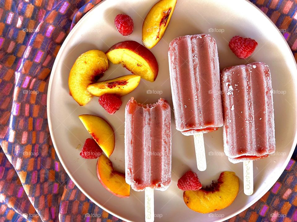 Three strawberry popsicles one with a bite taken with slices of nectarines and red raspberries on a pink plate lying on a purple orange and red patterned silk cloth