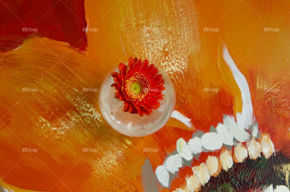 A single gerbera flower in a clear vase on a painting of gerbera flowers.