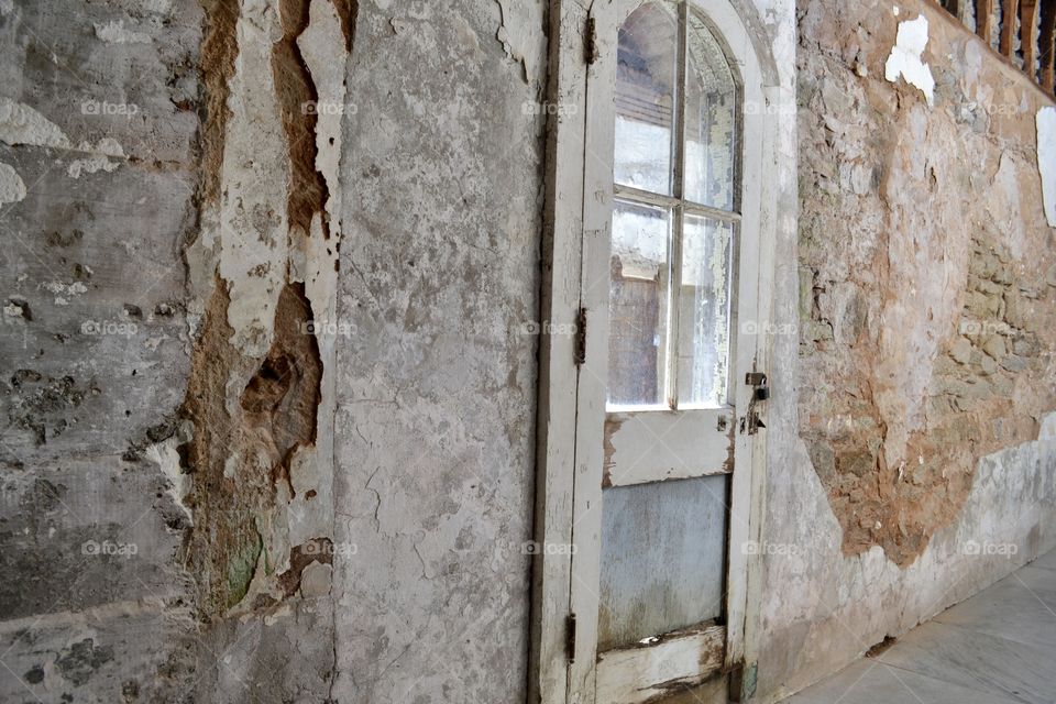 An old white wooden door with an arched window pane hanging in a decaying old wall with chipping paint