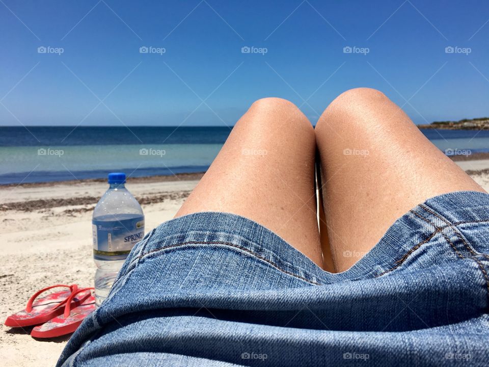 Woman sitting on beach, bare legs jean skirt water bottle to side toward ocean horizon