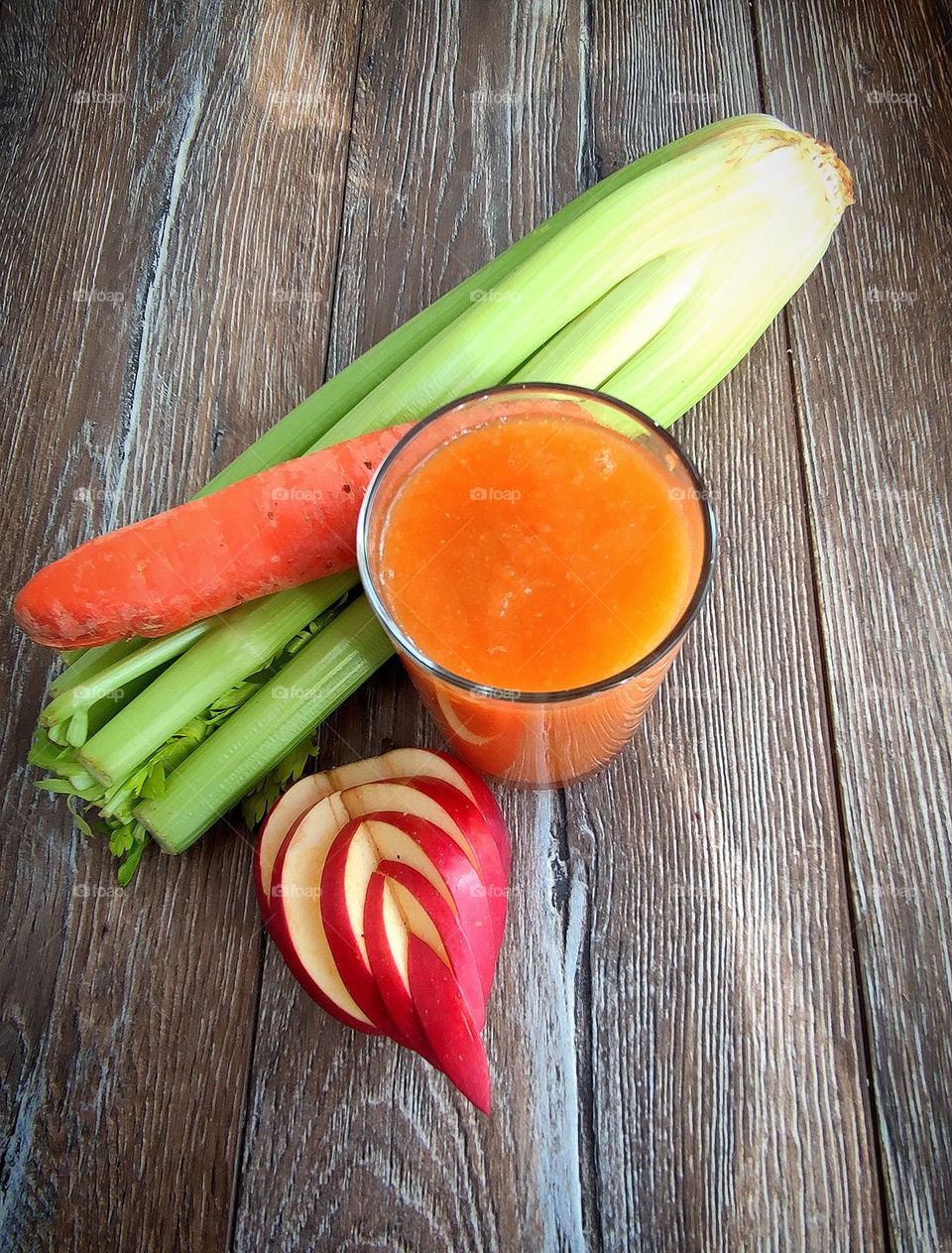 There is a glass with an orange smoothie on a wooden surface.  Ingredients lie nearby: a stalk of celery, a carrot and a red apple, which is cut into a heart shape