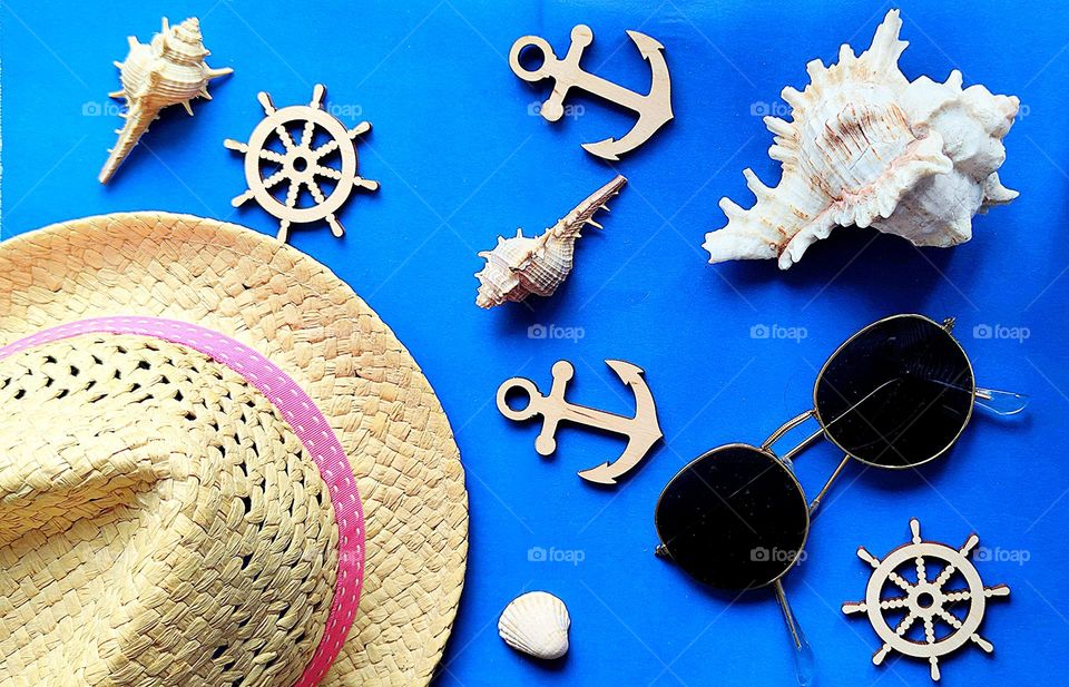 On a blue background are the essentials: a straw hat and sunglasses. Around are sea shells, wooden anchors and wooden helms