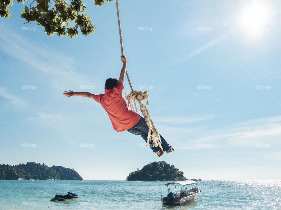 A boy hanging on a rope swing by the beach on a sunny day