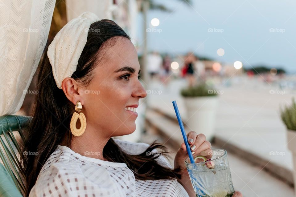 Side view of happy stylish young woman enjoying a cocktail in beach bar