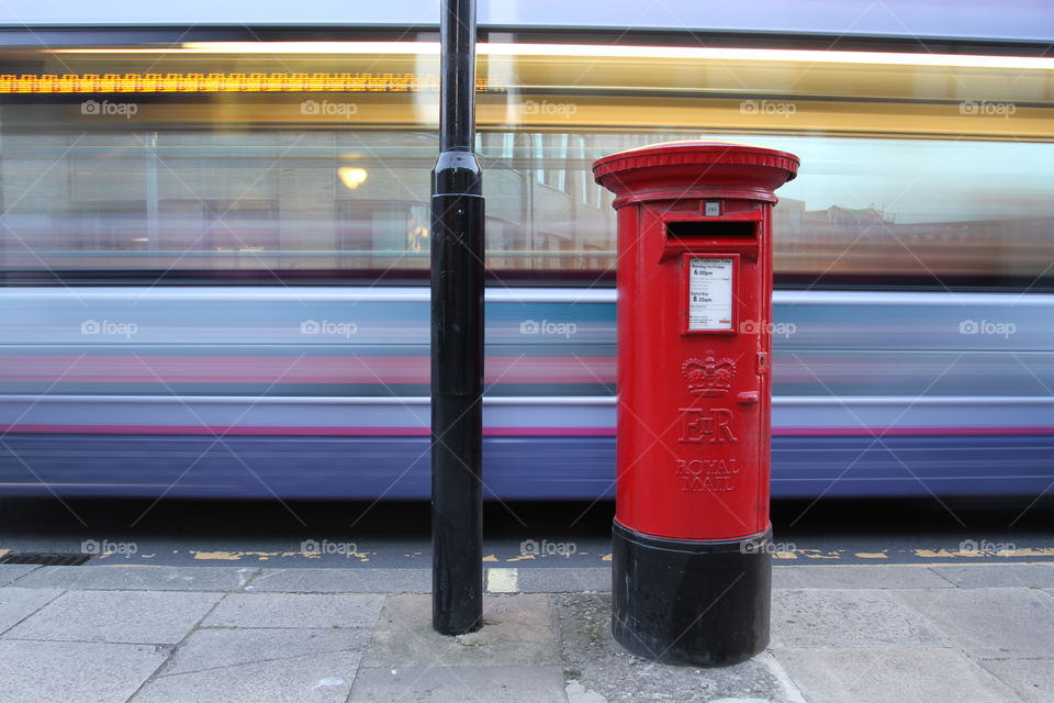 A bright red post box with a bus passing behind causing motion blur in the city of York