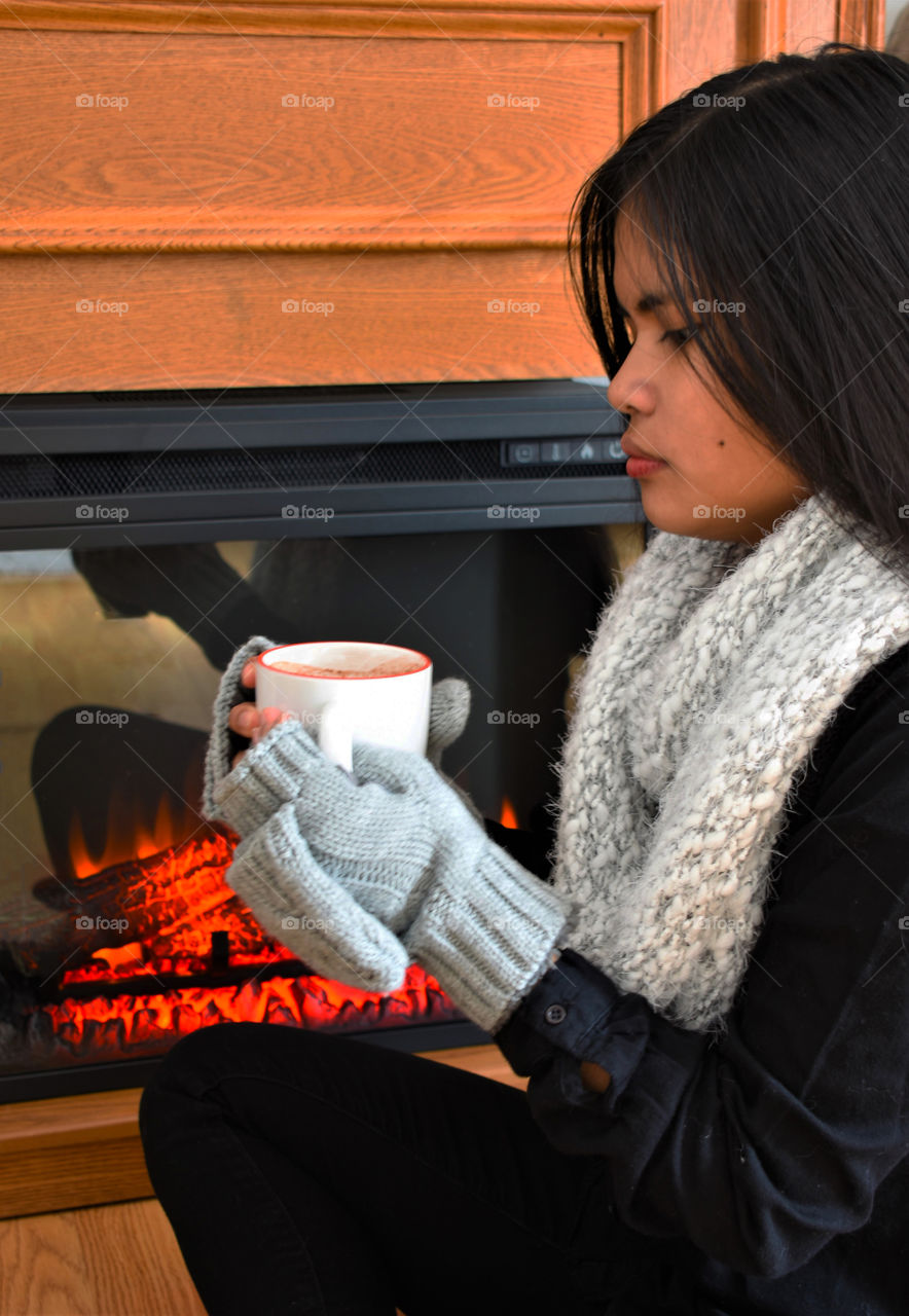 Girl beside fireplace with mittens and scarf on and holding a cup of hot cocoa 