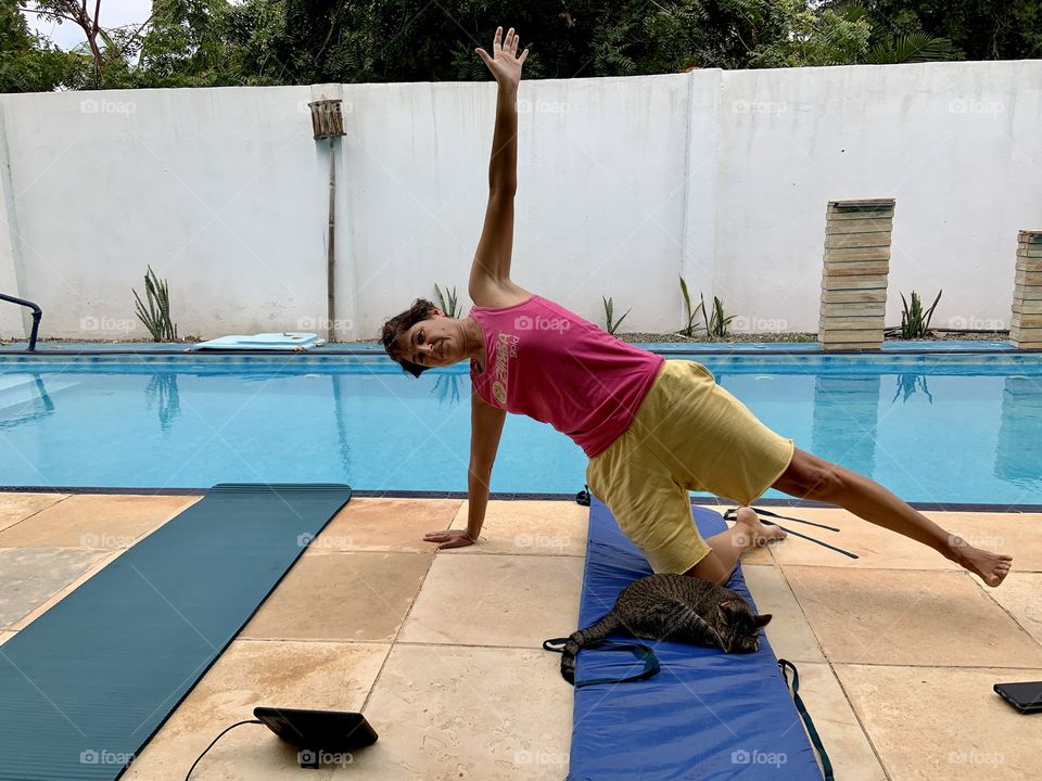 Woman playing yoga near swimming pool with cat on yoga mat 