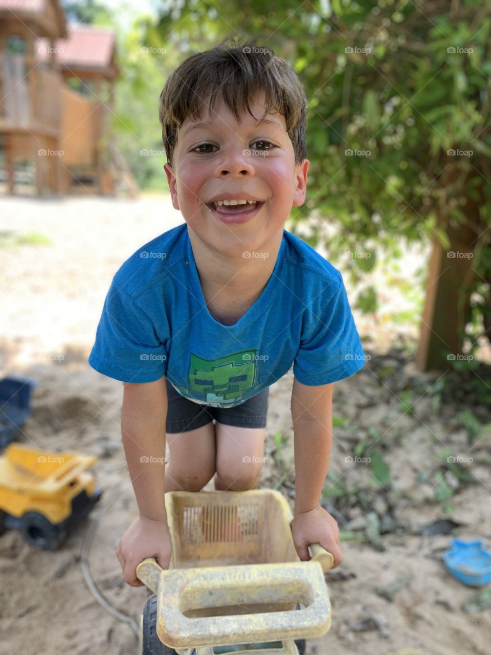 Sweaty, happy boy playing with a dump truck toy in a sandbox at a playground on a summer day.
