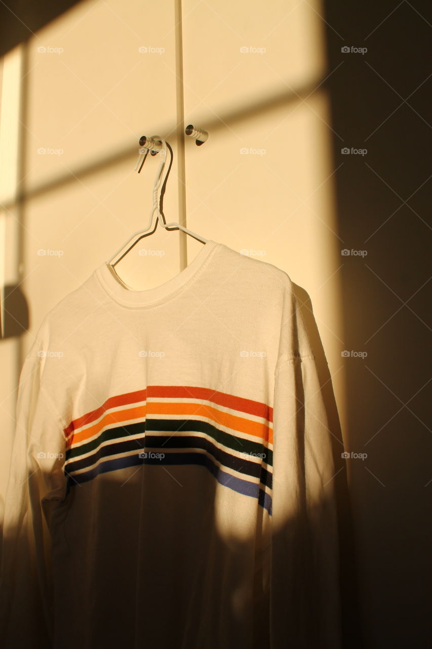 Rainbow sweater hanging on a white wall indoors with sunlight-no people