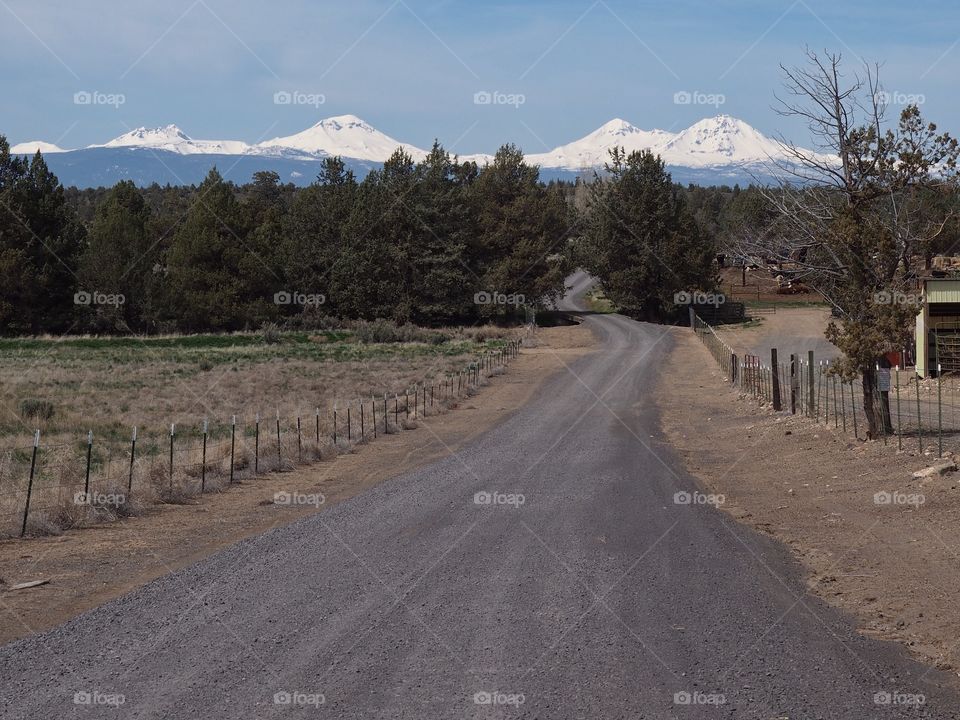 A gravel road winds through farmland with the snow covered Three Sisters in Oregon’s Cascade Mountain Range in the background in rural Central Oregon. 