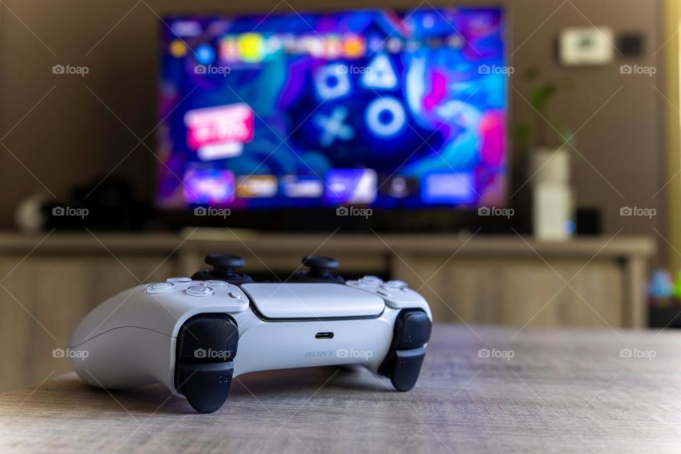 A portrait of a wireless ps5 controller lying in front of a television screen. the playstation 5 device is ready to play with.