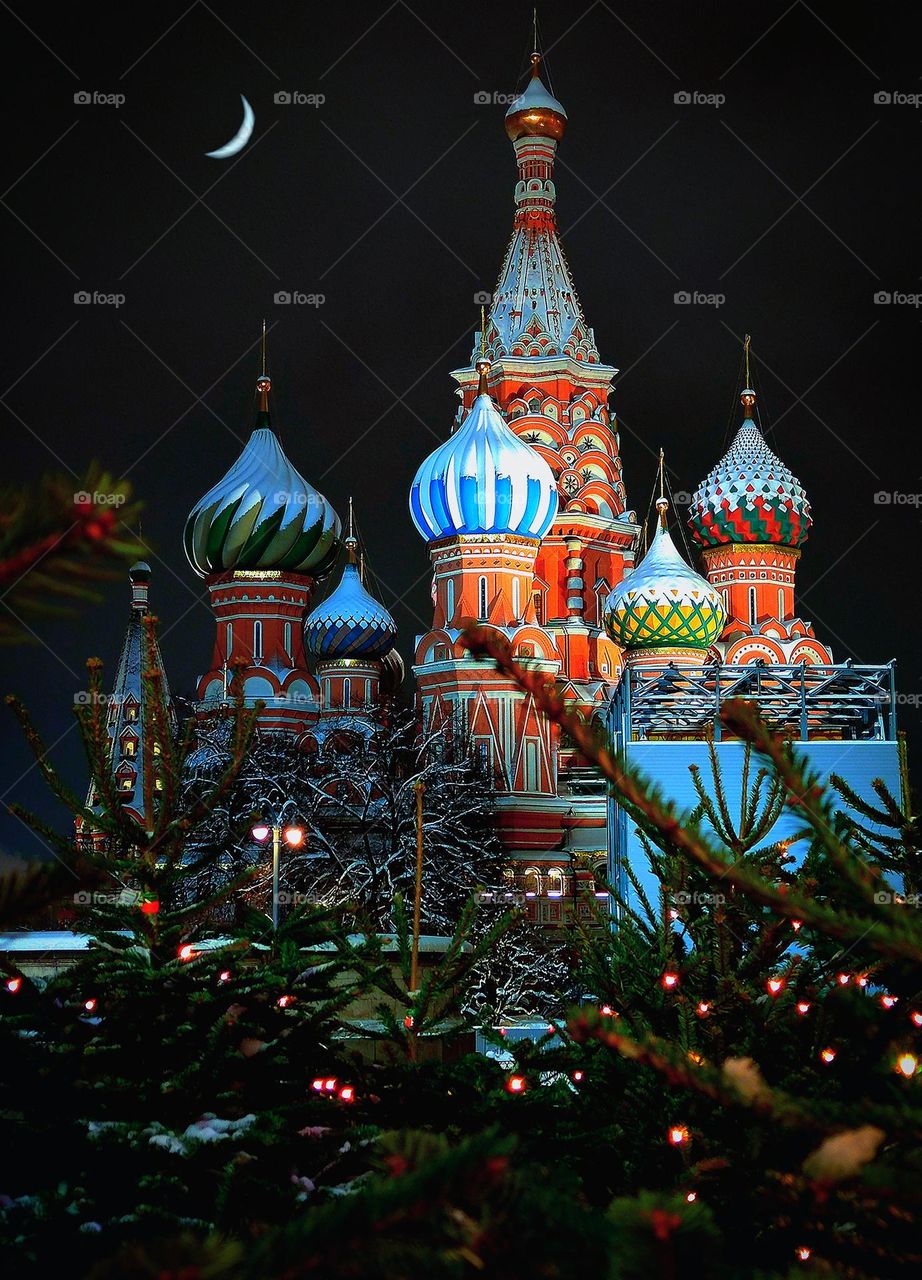 Winter fairy tale.  The multi-colored domes of St. Basil's Cathedral over which the sickle of the month shines.  There are Christmas trees in the snow near the temple.  In the foreground, branches of Christmas trees in luminous garlands. Moscow.