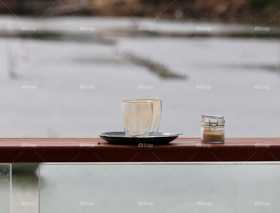 Two empty iced cappuccino glasses on black plate on ledge with mason jar of sugar on outdoor restaurant patio overlooking ocean, early morning, still life with copy text space