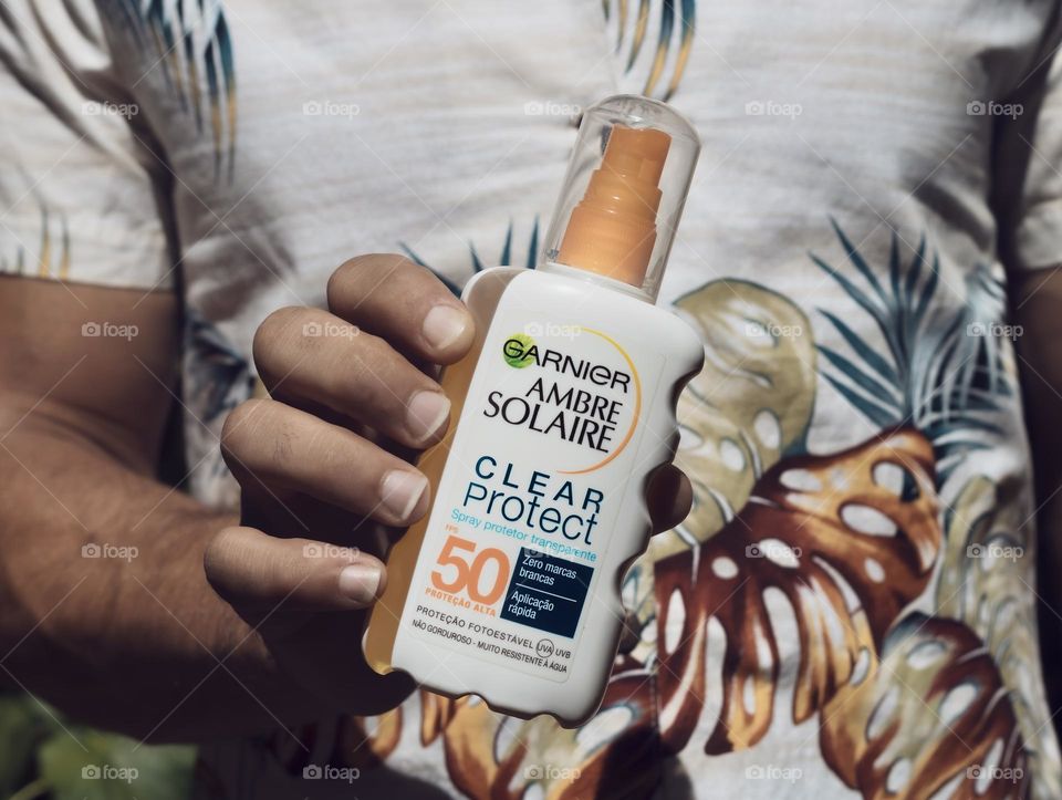 A tanned man in a vintage Hawaiian shirt holds out a bottle of sunscreen 