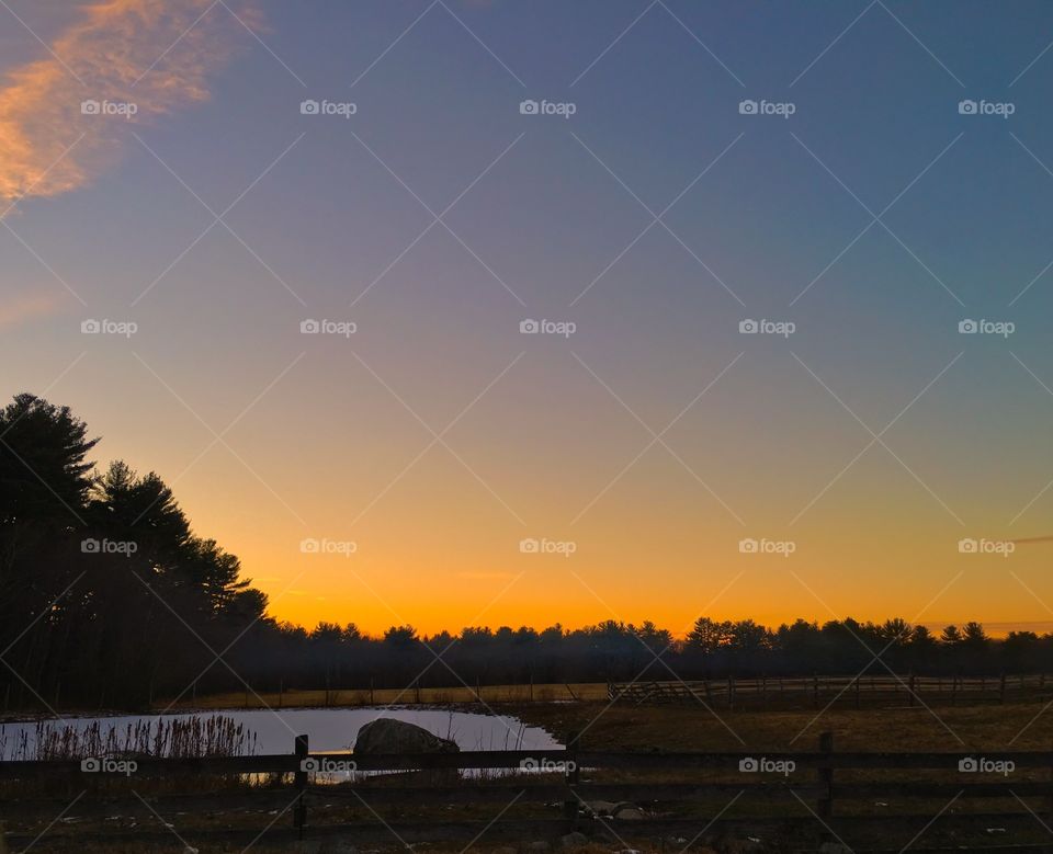 Rural landscape at sunset with dark sky and orange glow on the horizon reflected in the pond water, silhouetted trees in the background