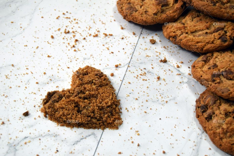 A heart made out of chocolate chip cookie crumbs sits alongside a pile of golden brown chocolate chip cookies on a white marble background.