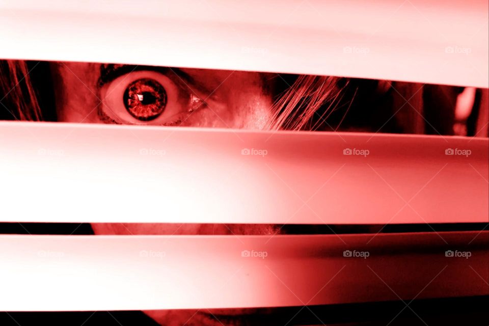 A wide-open eye looks through blinds in spooky red light 