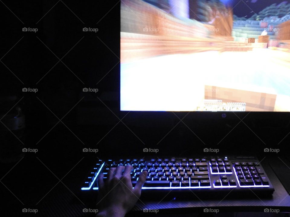 Teenager boy playing Minecraft on the gaming computer seen from his left side and close-up of his hand playing in a dark room.