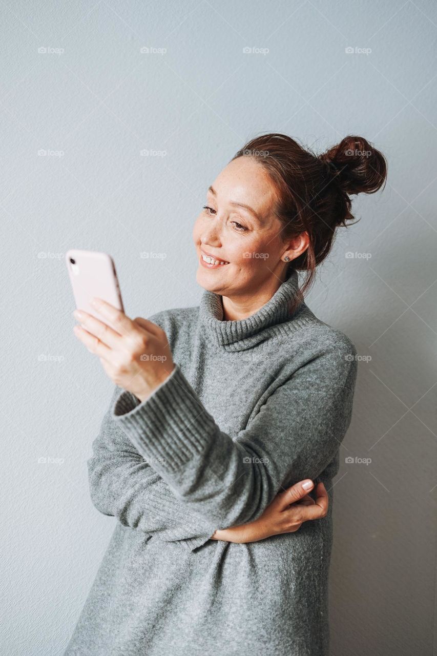 Vertical portrait of Adult beautiful smiling woman forty years with brunette curly hair in warm grey knitted dress using mobile on grey background