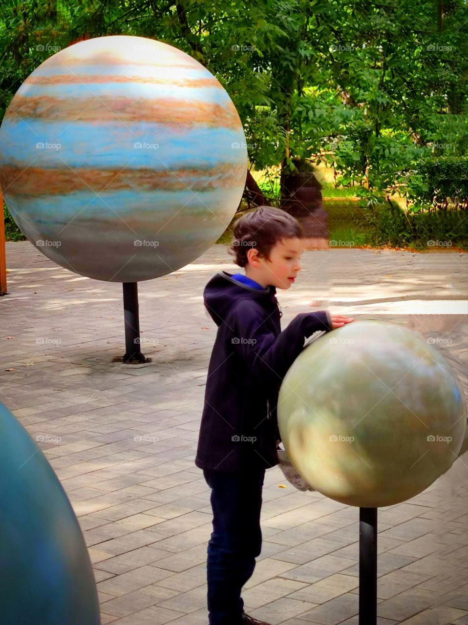 Children's playground among green trees. On the playground are round models of the planets. The boy looks at one of the planets with interest