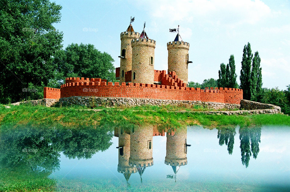 Fairy-tale like dreamy castle medieval red brick kids playground fortress reflected in water surface of the moat in children's park on a sunny summer day in Sumy, Ukraine