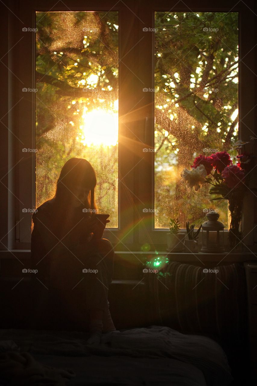 Little girl sitting with a phone in her hands by the window in the setting sun