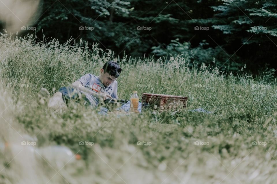 One young handsome caucasian man lies on a blanket with a wicker picnic basket, spread out food and drinks, watches a movie on a tablet in a meadow with tall grass on a summer day, close-up view from below.