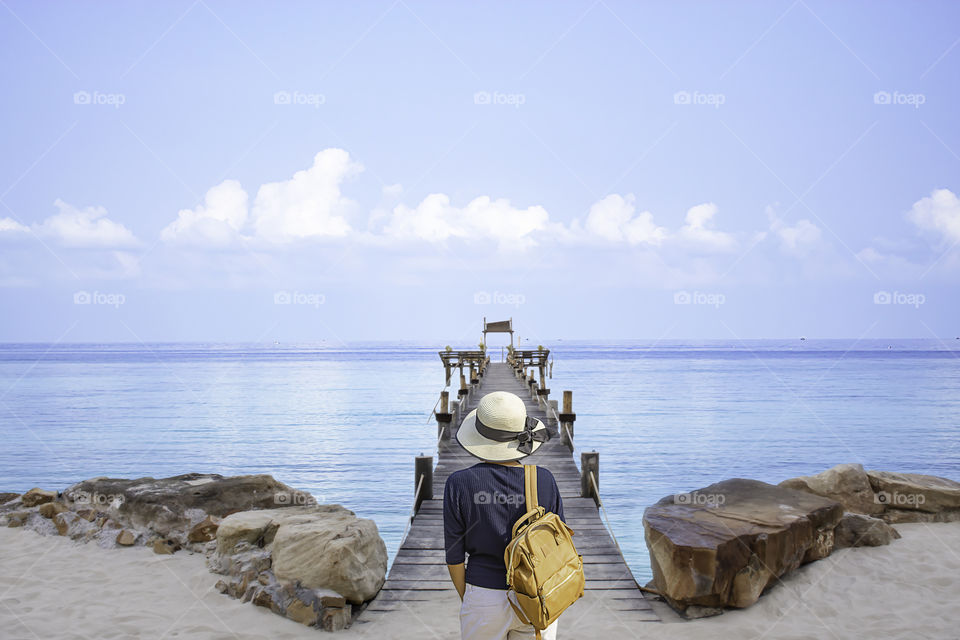 Women shoulder backpack and Wear a hat on wooden bridge pier boat in the sea and the bright sky at Koh Kood, Trat in Thailand.