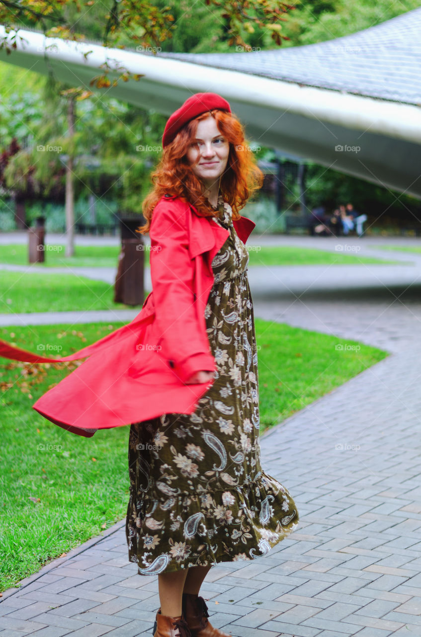 Young happy woman with red hair, freckles in a red beret and a coat walking in autumn park. Fall cold season