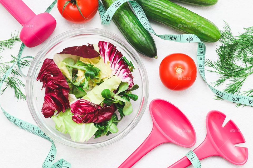 A glass bowl with fresh lettuce leaves, cucumbers, tomato, pink dumbbells, pink lettuce spoons and a green measuring tape lie on a light white cement background, flat lay close-up.