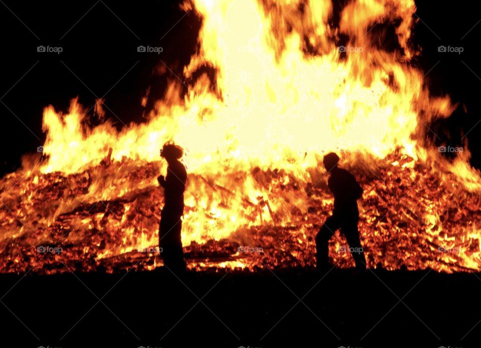 2 men are silhouetted against a huge roaring bonfire 