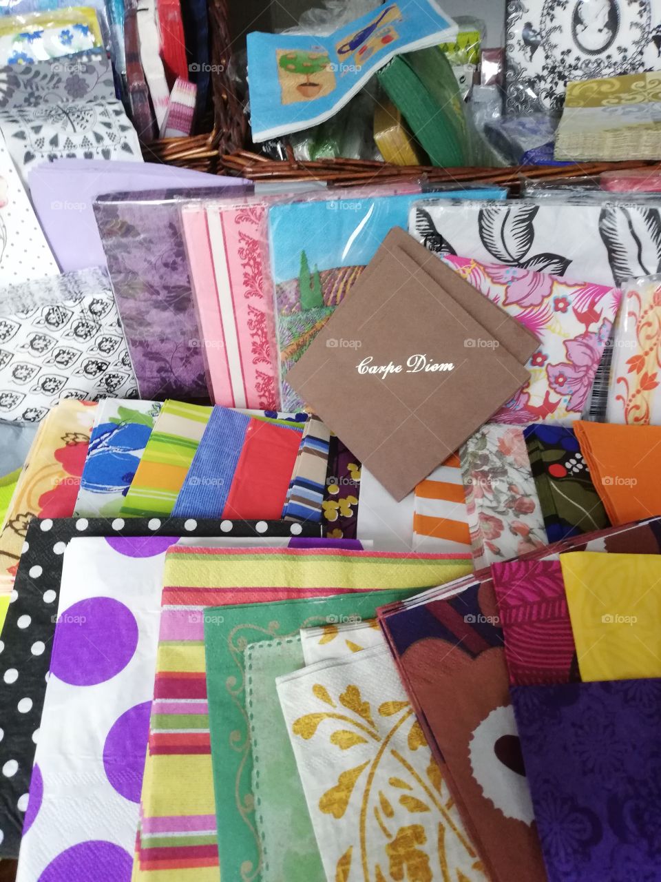 Different napkins in the plastic packages in the brown rattan basket, some are on the table one on the other. Many colours, figures, spots, drawings. In the front the text carpe diem on the brown napkin with golden letters.